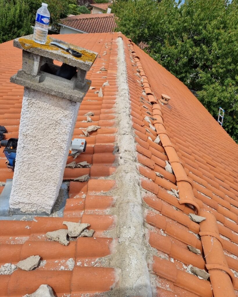 Toit en tuiles orange, cheminée et faîte avec débris de ciment. Une bouteille d'eau et un outil sur la cheminée. Travaux de réparation en cours.