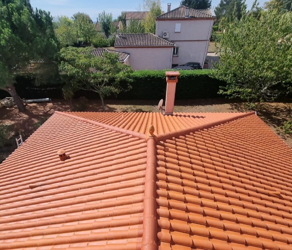 Toit de maison en tuiles de terre cuite orangées sous le soleil, avec un faîtage élaboré, un finial et une cheminée. Arbres et habitations en fond.