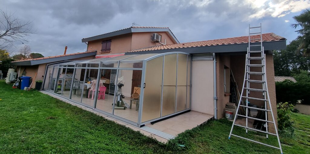 Véranda en verre et aluminium adossée à une maison aux tuiles oranges. Un jardin verdoyant et une échelle sont visibles.