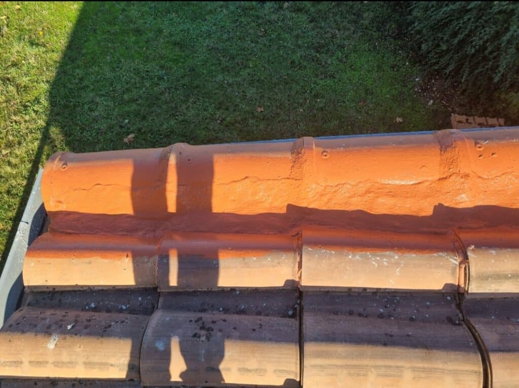 Vue rapprochée de tuiles faîtières de toit orange avec fissures et partie rénovée, ombres solaires sur la surface, pelouse.
