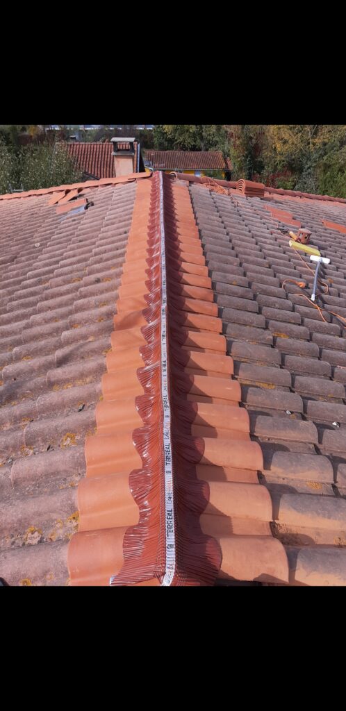 Rénovation d'un toit en tuiles terracotta avec installation de faîtières neuves. Bande de fixation TERREAL visible sur le faîtage. Outils de couvreur sur la droite.