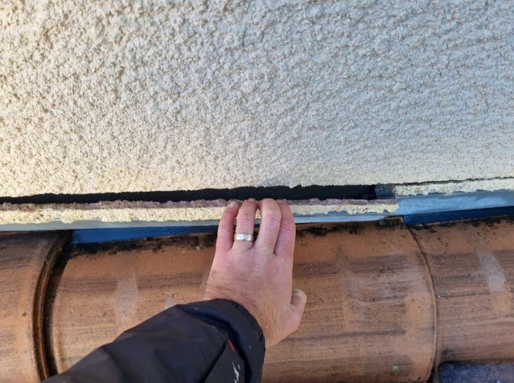 Problème d'étanchéité mur-toit : Écart visible à inspecter Main inspectant un large interstice entre un mur crépi et des tuiles de toit en terre cuite, suggérant un défaut d'étanchéité.